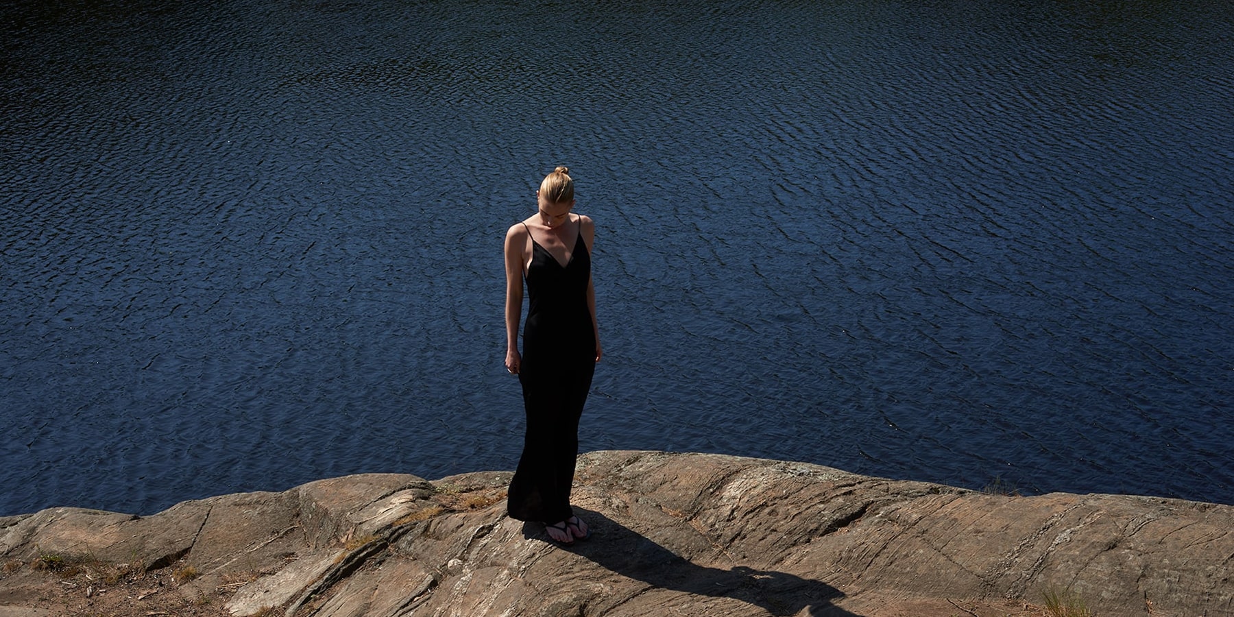 Woman in black long dress standing on a rock next to blue water.