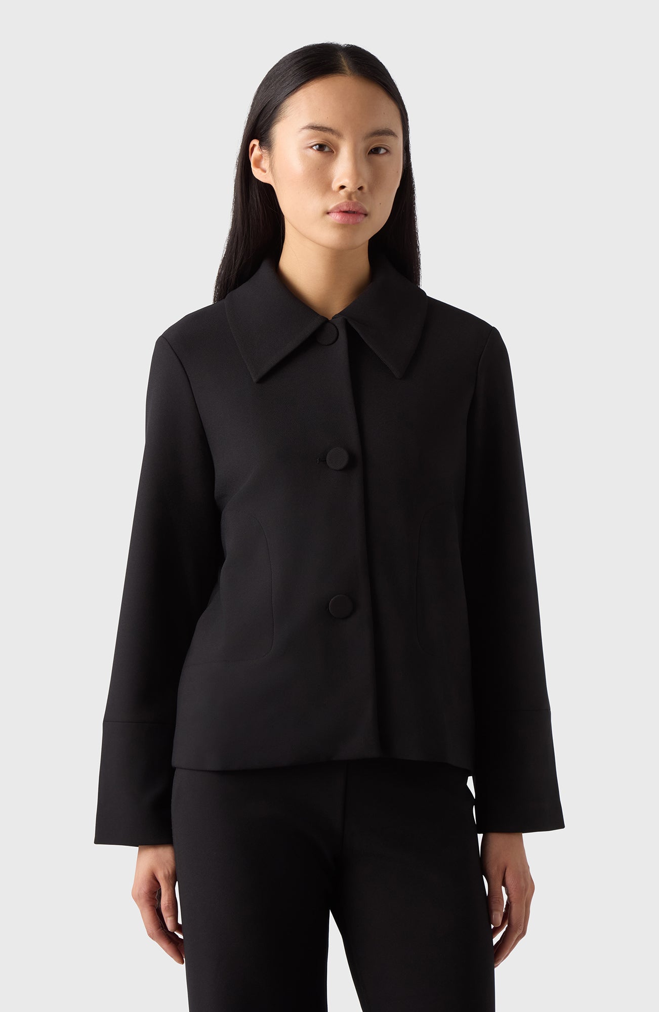 A woman standing against a white background wearing a navy blazer with a classic collar and tailored cut, paired with blue jeans and beige sandals.