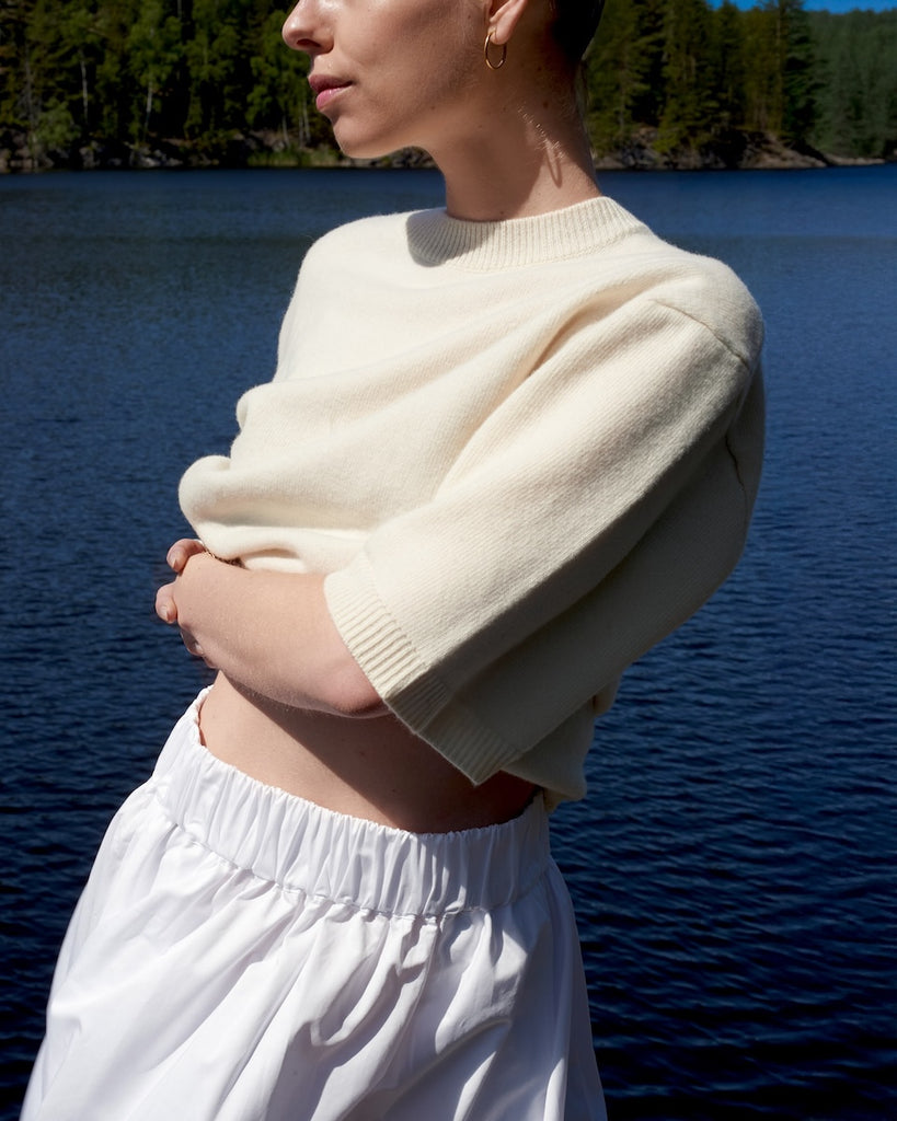 Person wearing a cream sweater and white pants standing by a lake with trees in the background
